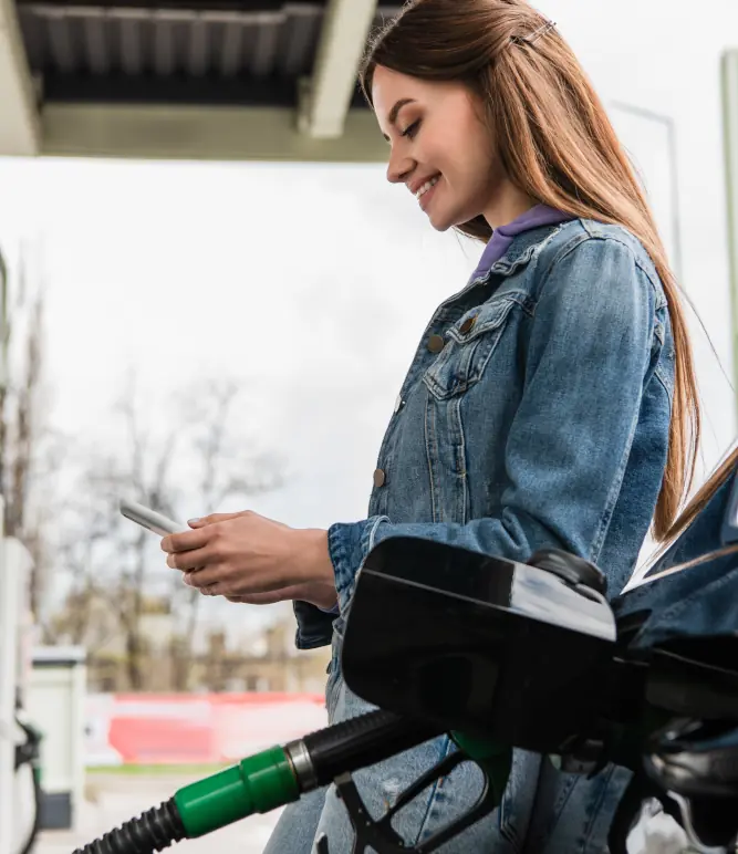 Frau steht an Tankstelle bei einer Zapfsäule - tankt und benutzt ihr Mobiltelefon